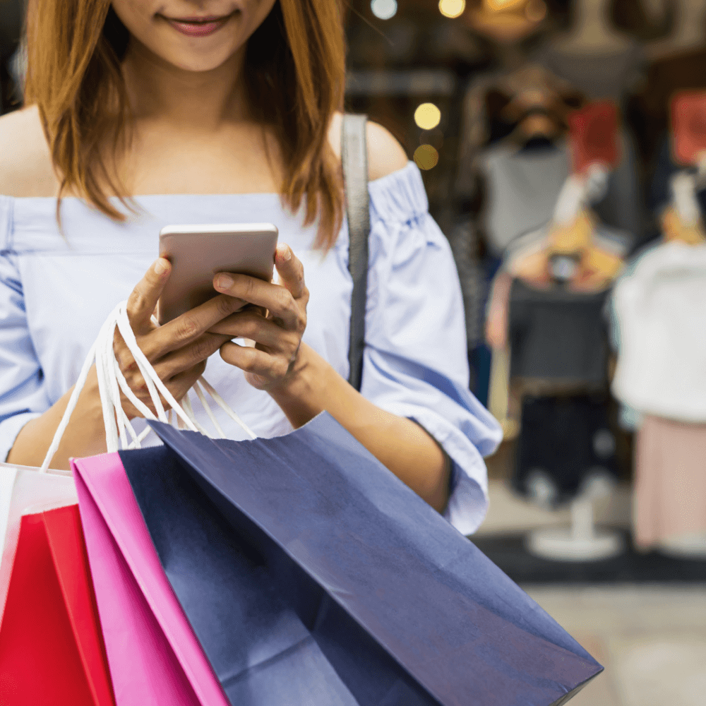 A woman holding shopping bags in one hand while using her phone with the other, standing outdoors near shops. - Home Instead