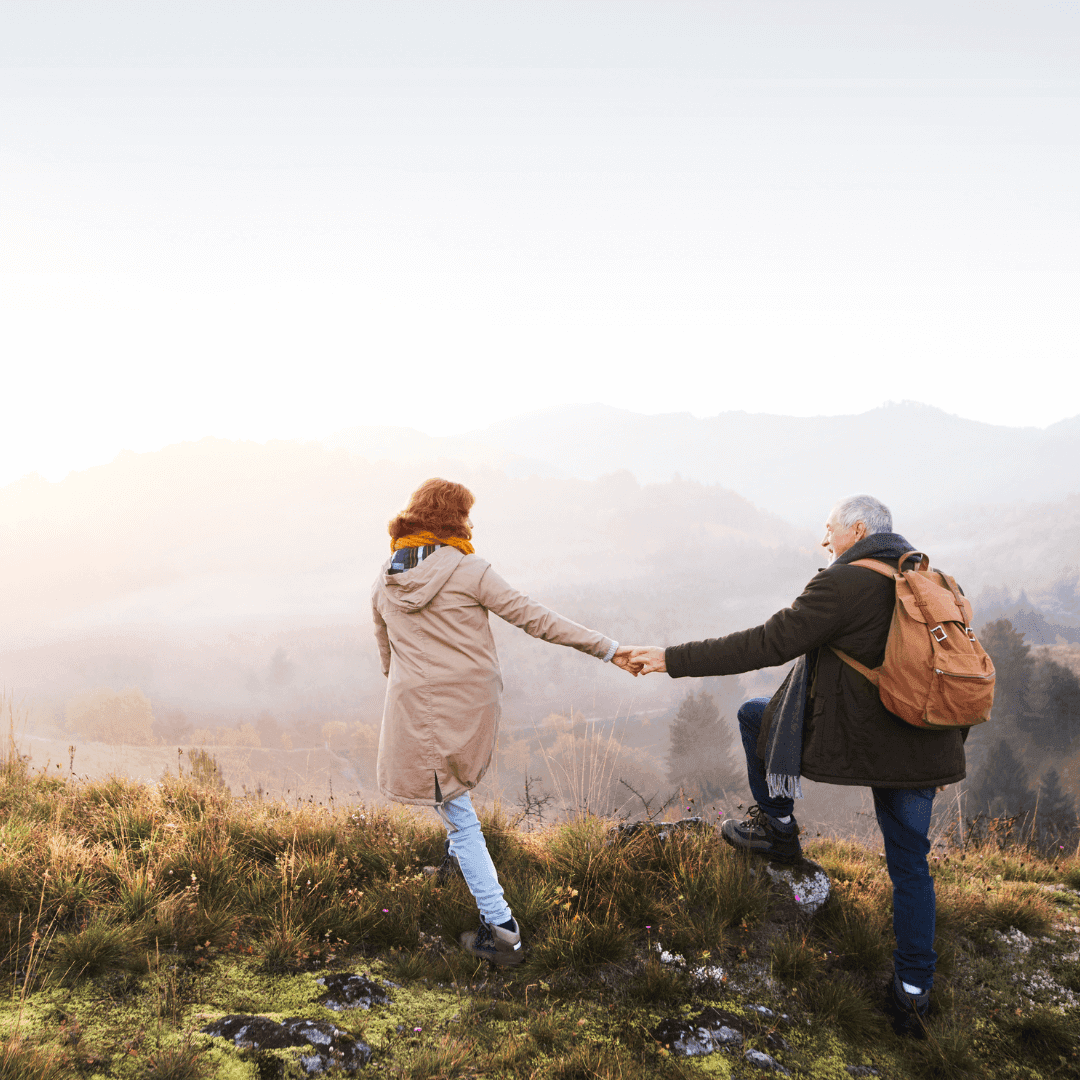 A couple holding hands while hiking on a grassy hilltop with a misty mountain landscape in the background. - Home Instead