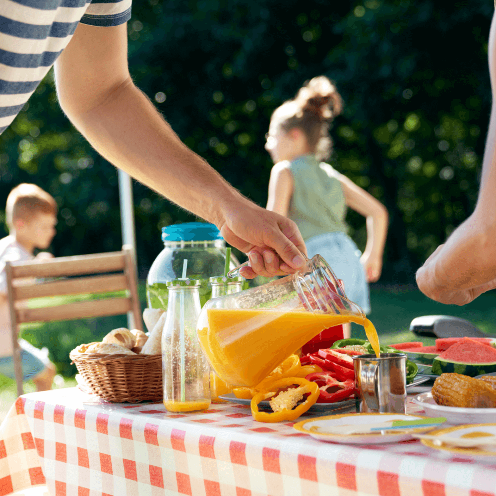 Person pouring orange juice at an outdoor picnic, with children playing in the background. - Home Instead