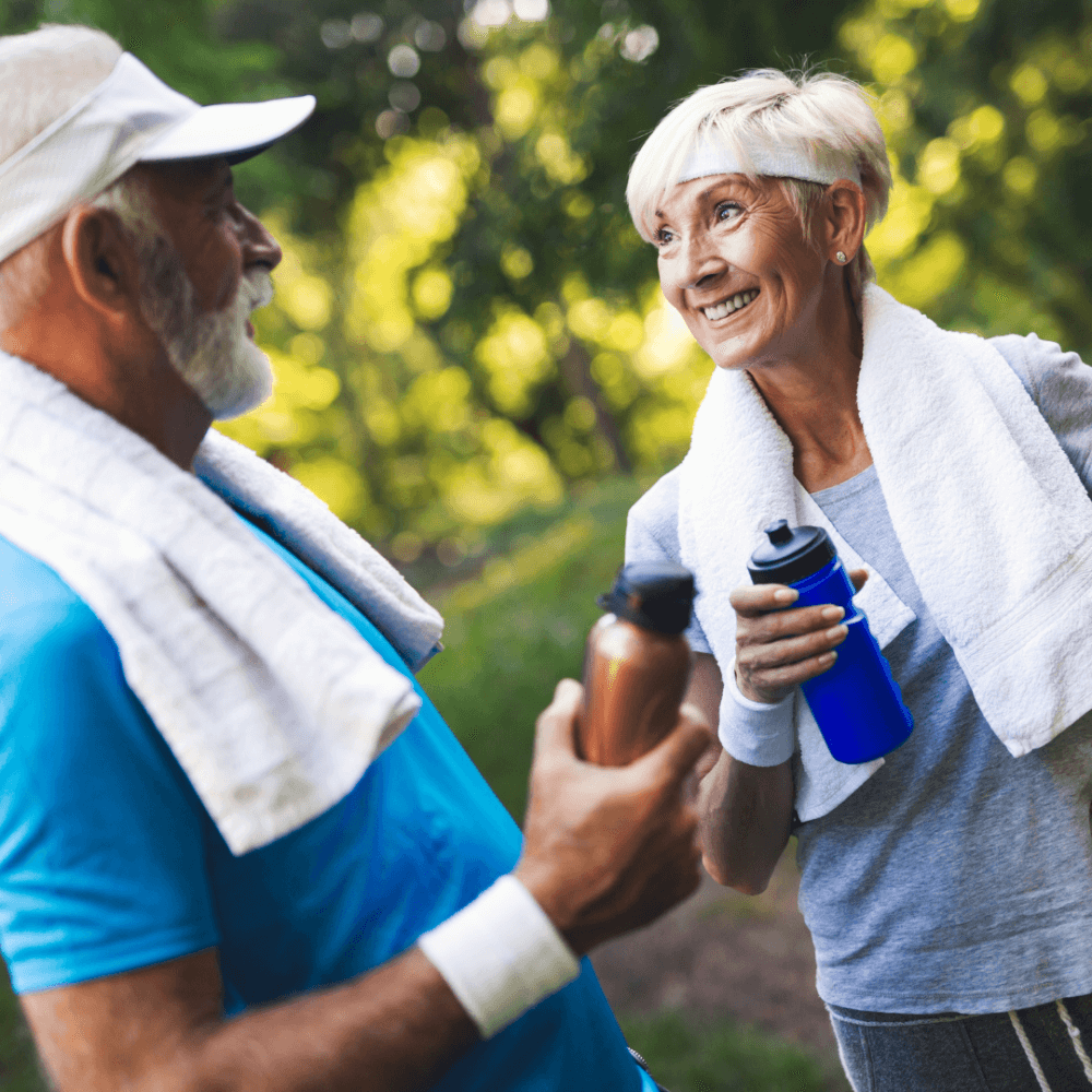 Two elderly people smiling at each other while holding water bottles and wearing workout gear and towels around their necks. - Home Instead