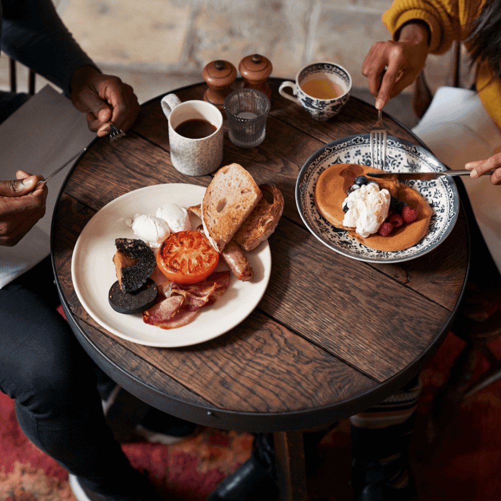 Two people eating breakfast at a rustic wooden table, featuring toast, poached eggs, bacon, and pancakes with fruit. - Home Instead