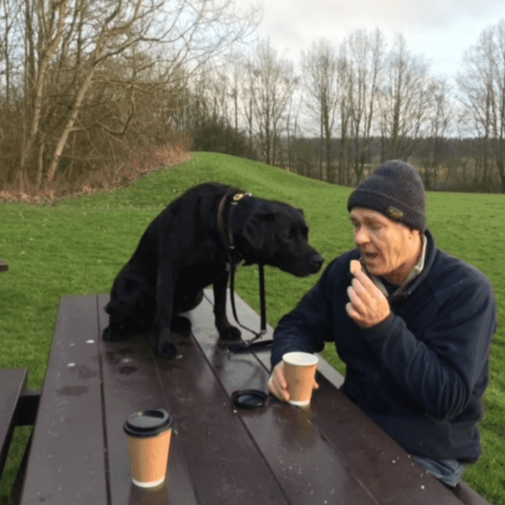 Man sitting at a picnic table with a black dog, drinking coffee outside on a grassy field with trees in the background. - Home Instead