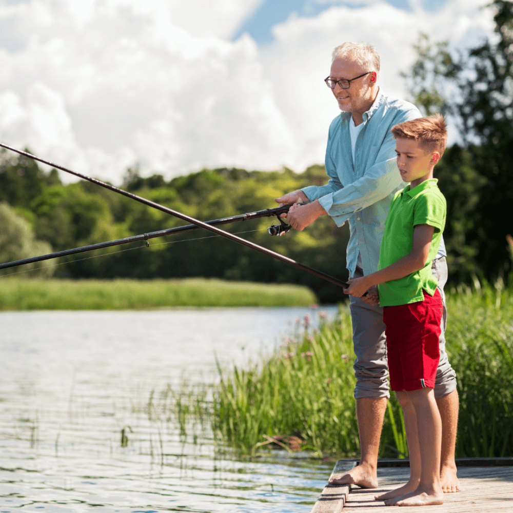 An older man and a young boy are fishing together on a wooden dock by a lake, surrounded by lush greenery. - Home Instead
