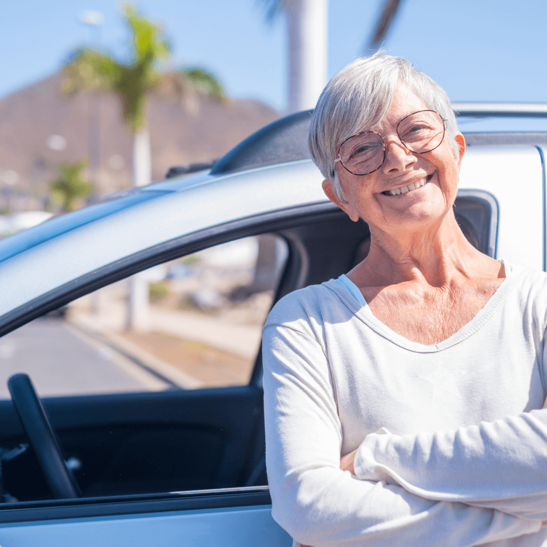 Smiling older woman with short gray hair stands with arms crossed next to a car on a sunny day. - Home Instead