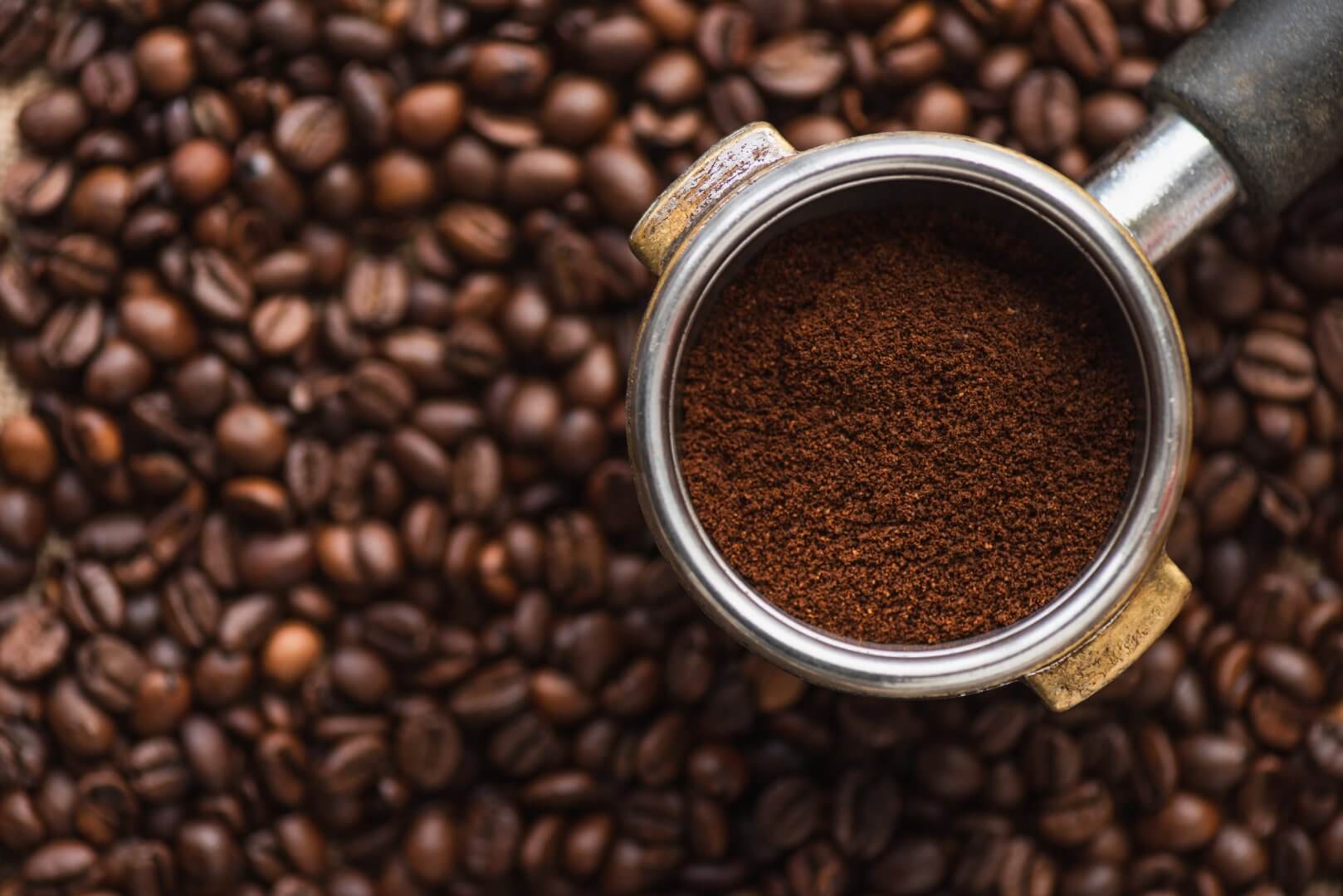 A portafilter filled with ground coffee placed on a bed of whole coffee beans. - Home Instead