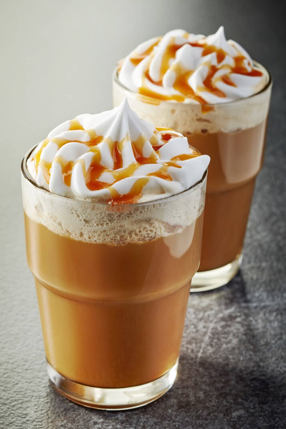 Two glasses of caramel macchiato topped with whipped cream and caramel drizzle on a textured surface. - Home Instead