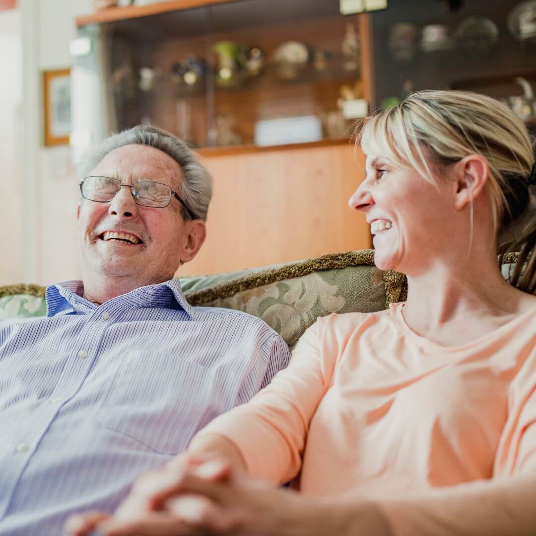 An elderly man and a woman sitting on a couch, laughing and smiling together, with shelves and decor in the background. - Home Instead