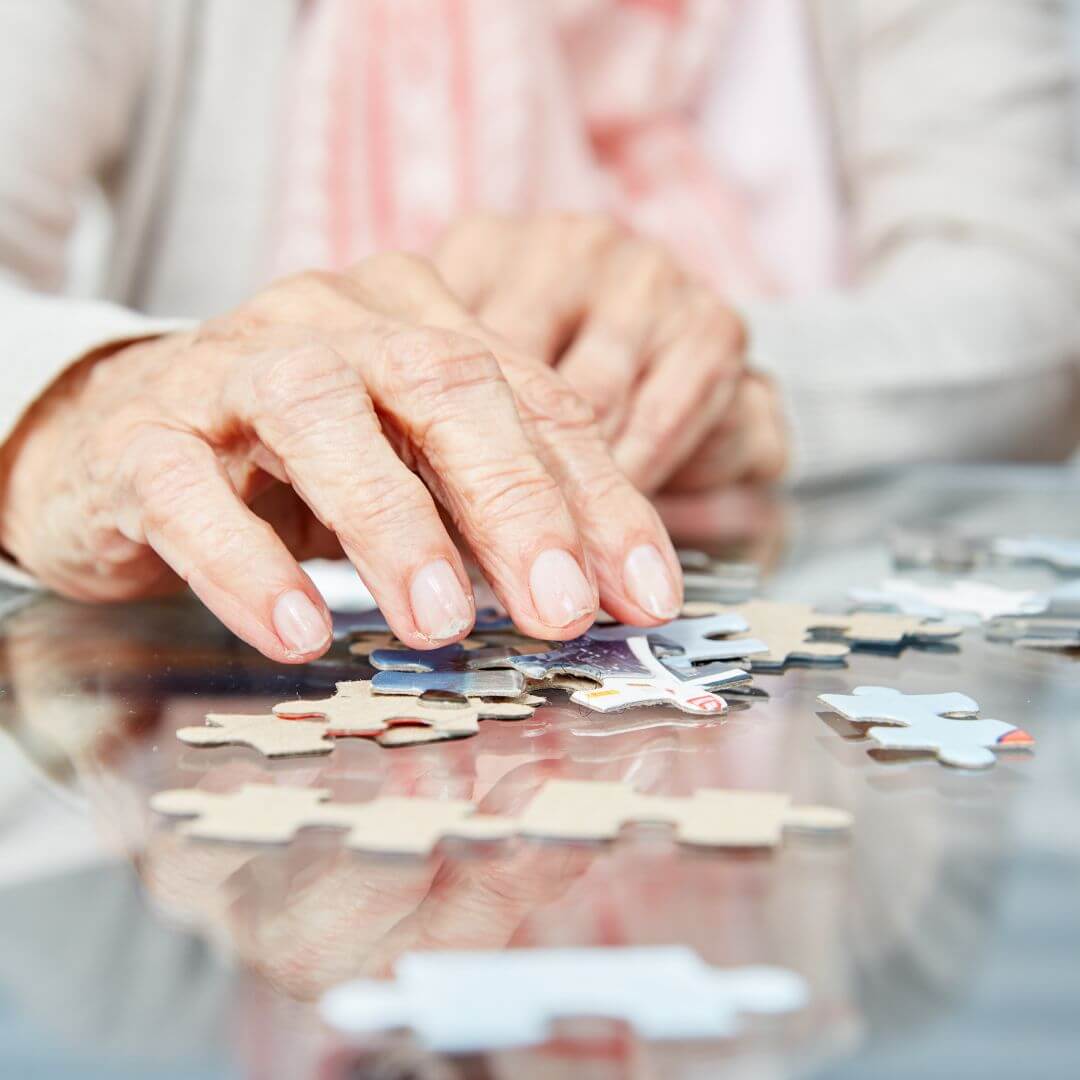 Close-up of an elderly person's hands assembling a jigsaw puzzle on a glass surface. - Home Instead