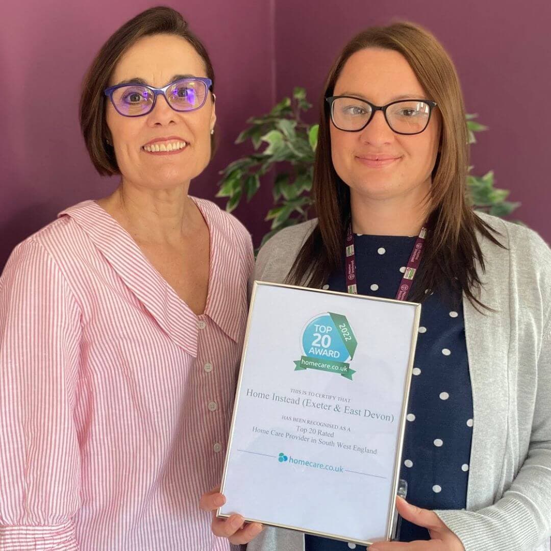Two women smiling and holding a framed award for Home Instead, recognizing them as a top home care provider in South West England. - Home Instead