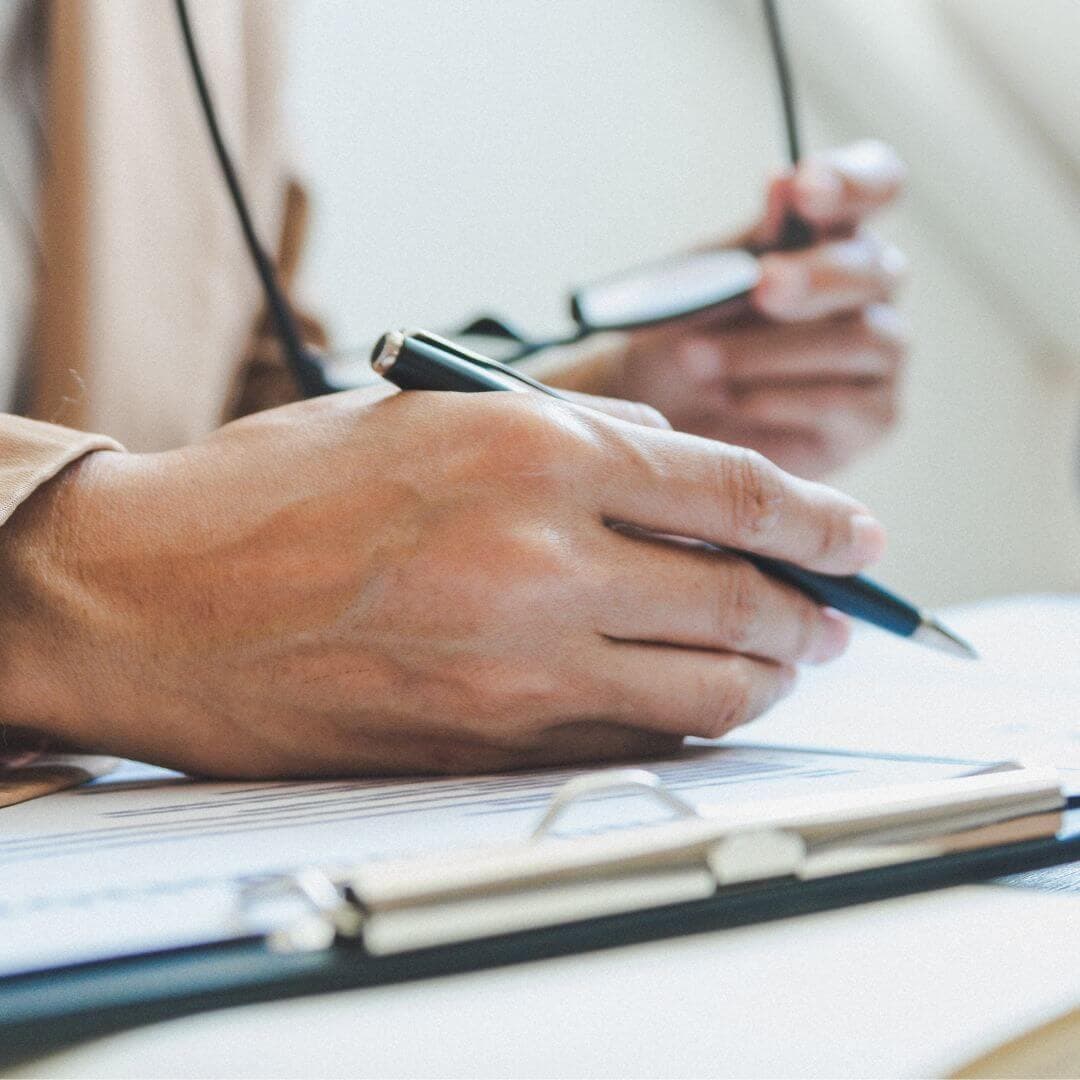 A person holding a pen and adjusting glasses while working on documents on a clipboard. - Home Instead