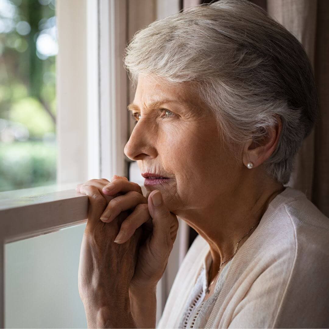 Elderly woman with short gray hair gazing out of a window, lost in thought, with her hands clasped together near her chin. - Home Instead
