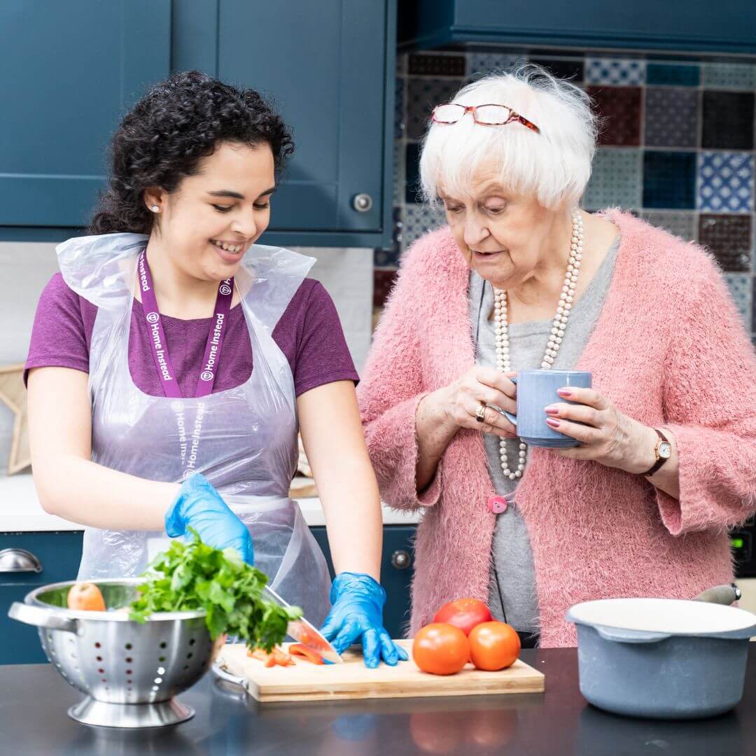 A caregiver in gloves helps an elderly woman in the kitchen, preparing vegetables while the elderly woman holds a mug. - Home Instead