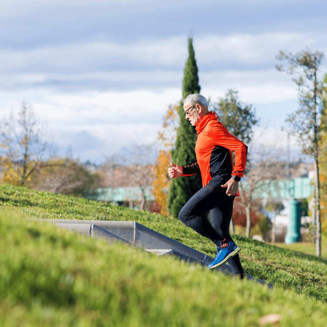 Elderly man in red jacket running uphill on a grassy slope with trees and cloudy sky in the background. - Home Instead
