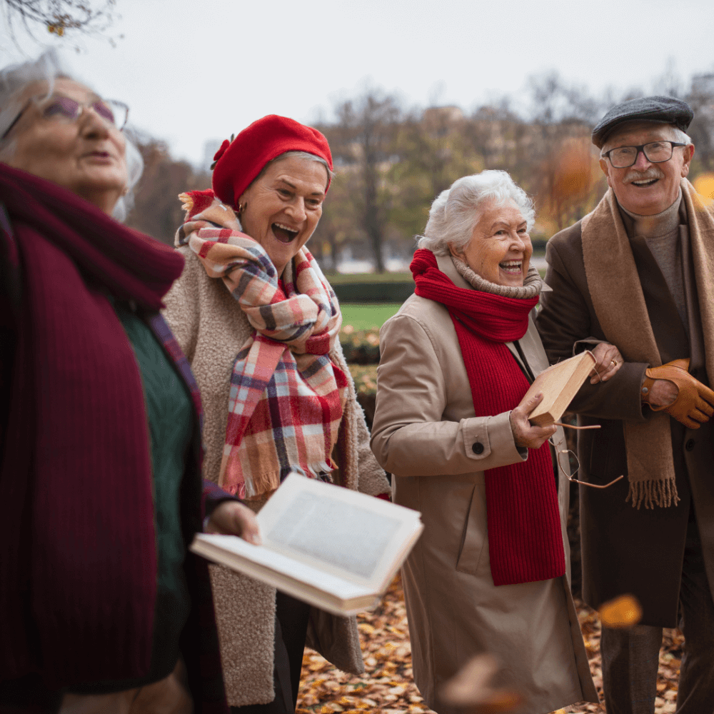 Four elderly people in warm clothes, laughing and holding books, standing in a park with autumn foliage. - Home Instead
