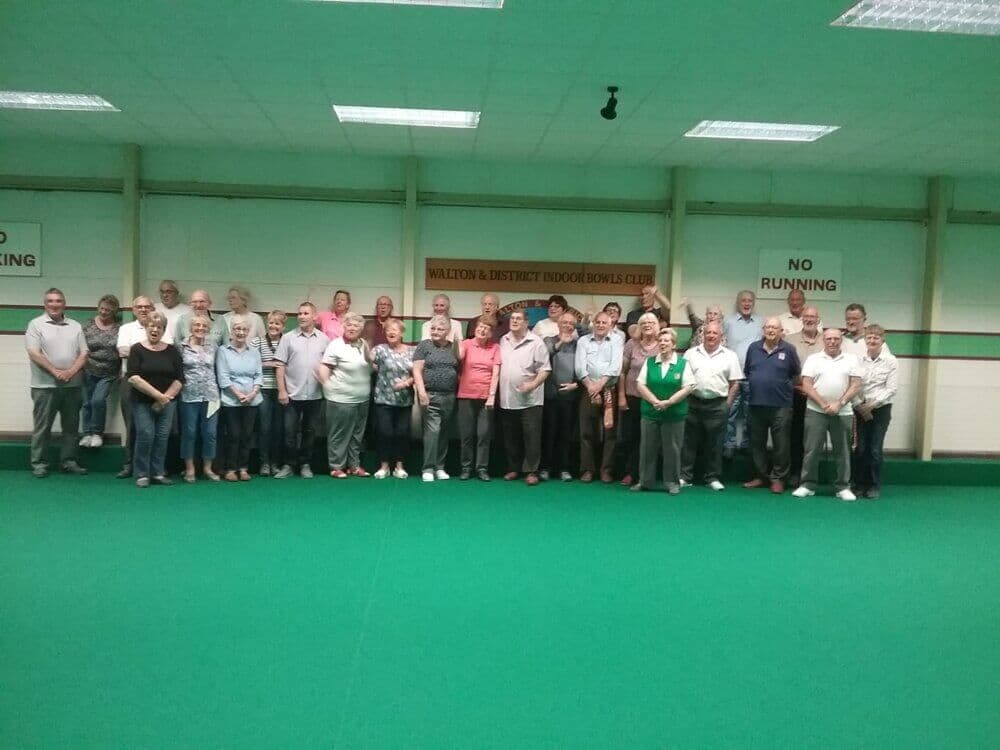 A group of people standing on a green indoor bowling field at Walton & District Indoor Bowls Club. - Home Instead
