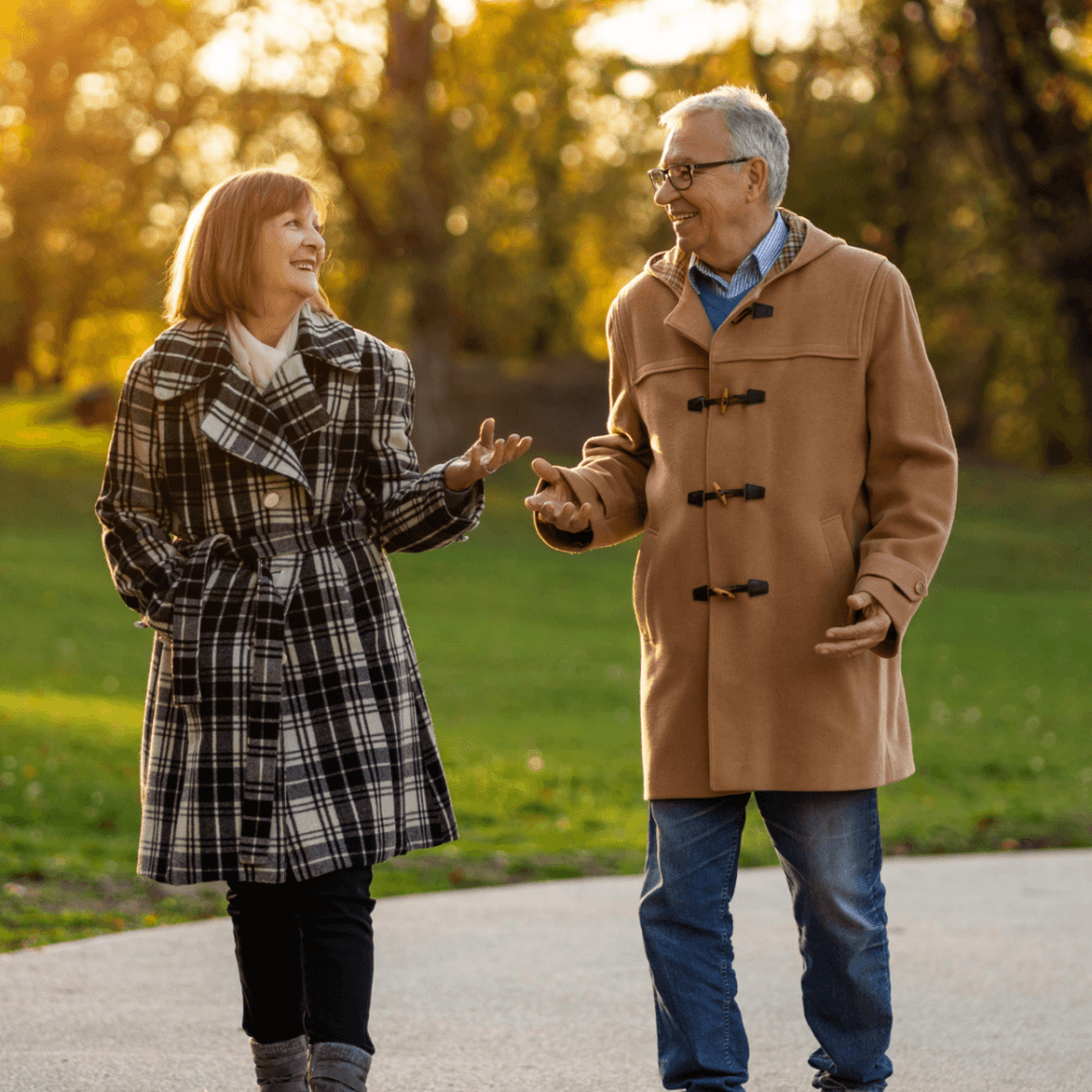 Older couple in coats walking and talking on a park pathway during a sunny autumn day. - Home Instead