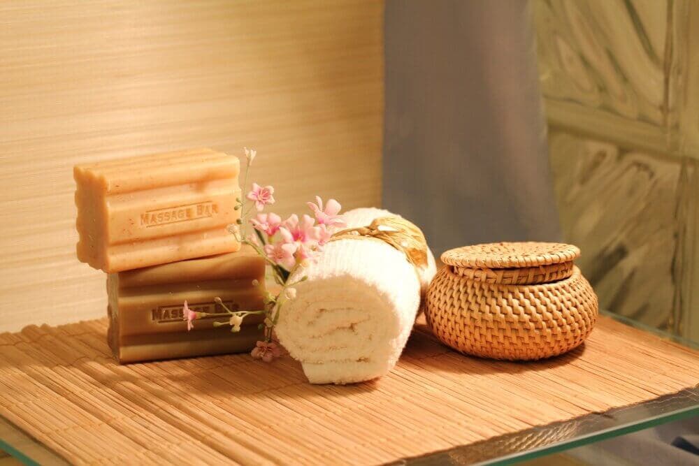 A spa setup with soap bars, a rolled towel, small flowers, and a wicker container on a bamboo mat. - Home Instead