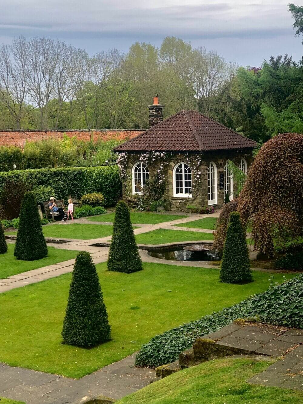 Stone cottage with a red roof in a garden with manicured shrubs, a pond, and a couple sitting on a bench. - Home Instead