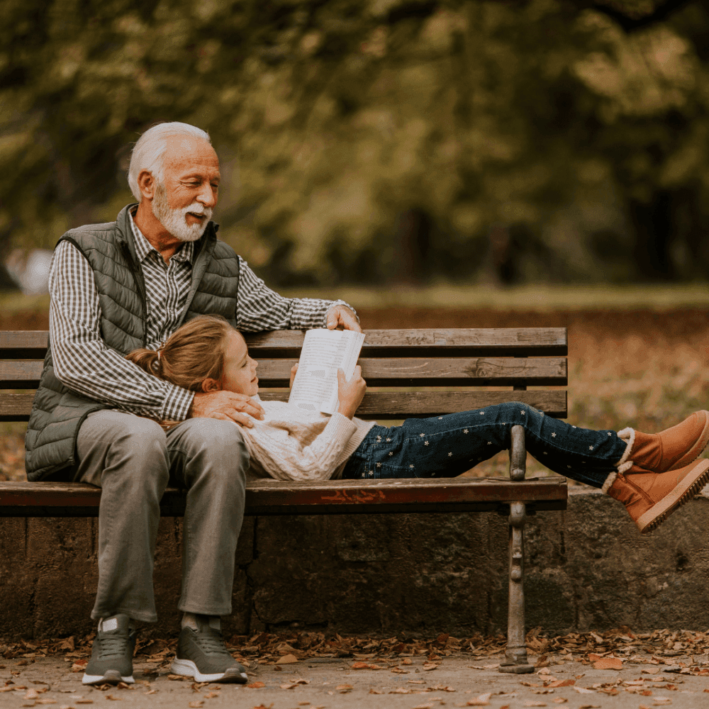 An elderly man and a child reading a book on a park bench surrounded by fall foliage. - Home Instead