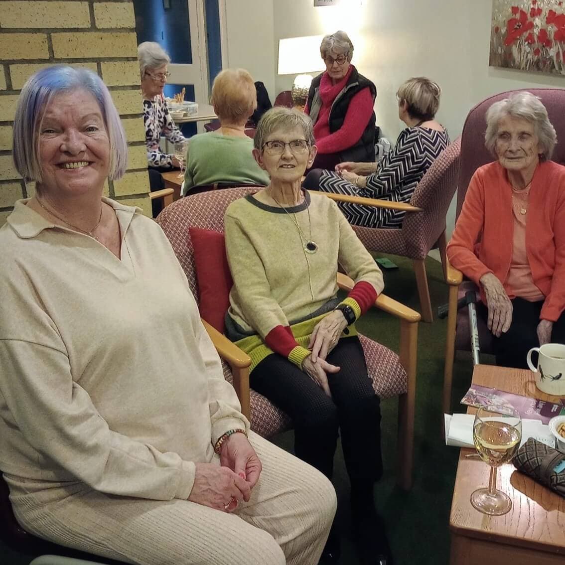 A group of elderly women sitting and talking in a cozy room with drinks and snacks on the tables. - Home Instead