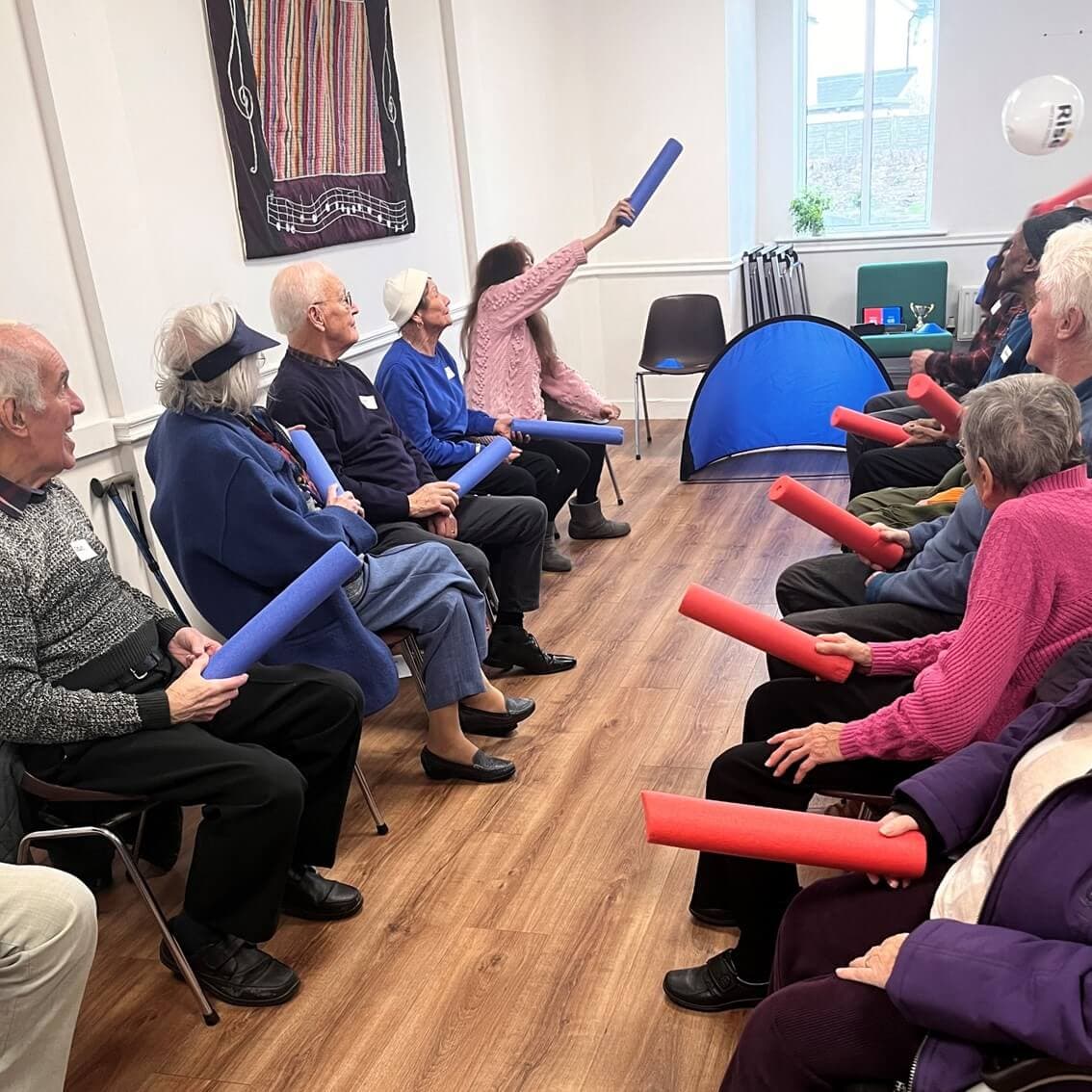A group of elderly people sitting in a circle, playing with foam noodles and a beach ball in a brightly lit room. - Home Instead