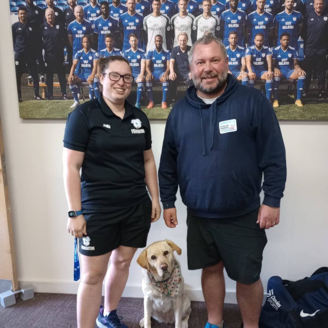 Two people smiling with a dog indoors, standing in front of a team photo hanging on the wall. The dog sits between them. - Home Instead