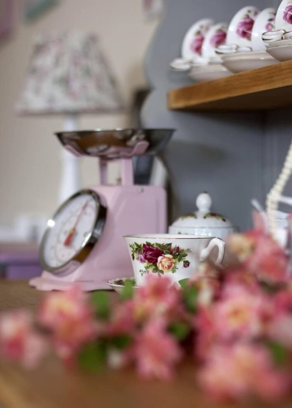 Floral teacups and a pink vintage kitchen scale on a wooden counter with blurred flowers in the foreground. - Home Instead
