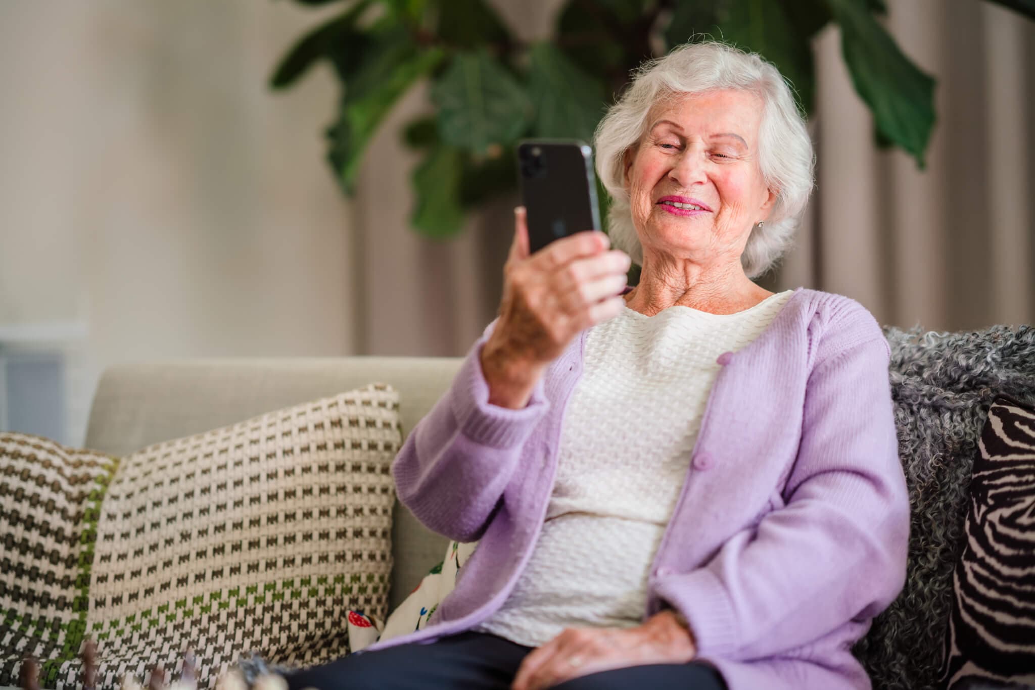 An elderly woman sits on a sofa, smiling while looking at her smartphone, with plants and pillows in the background. - Home Instead