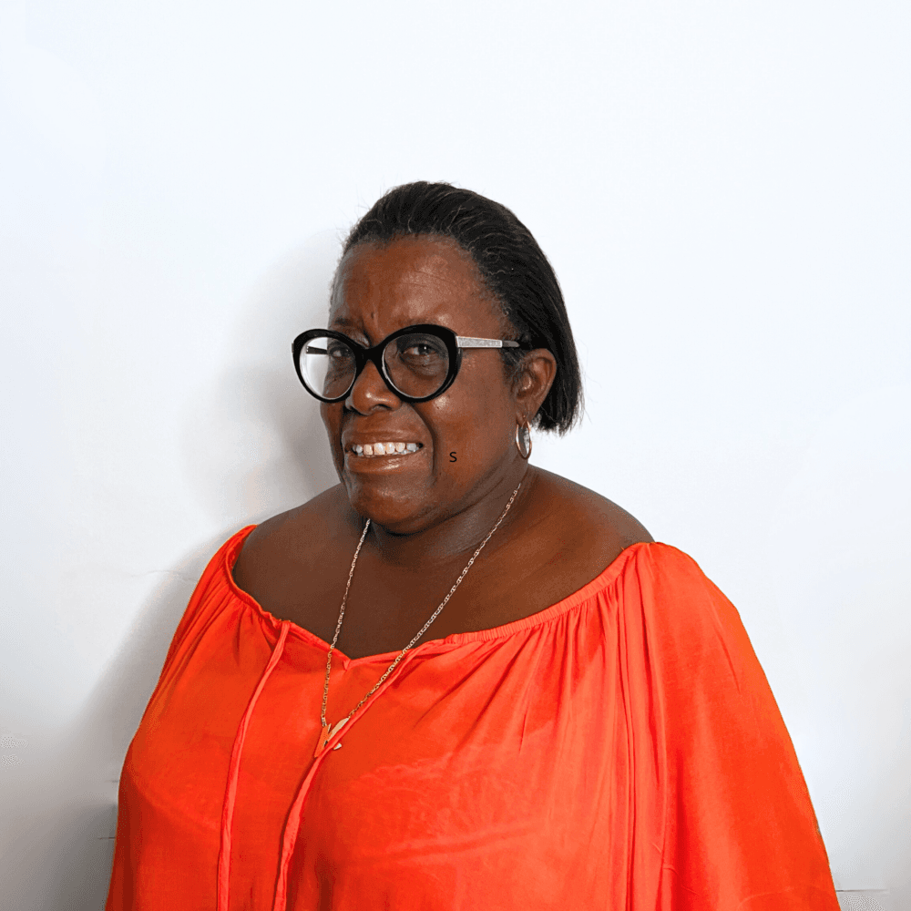 A person wearing a bright red off-shoulder top and round black glasses, smiling against a white background. - Home Instead
