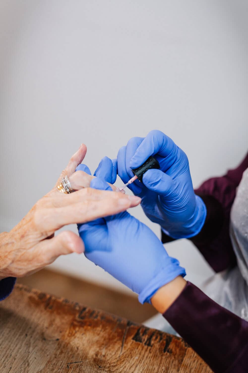 Close-up of a person wearing blue gloves applying nail polish to another person's fingernails on a wooden surface. - Home Instead