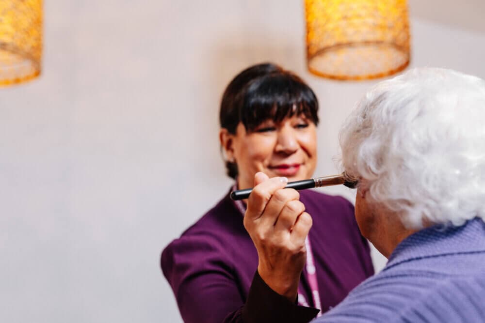 A woman applying makeup to an elderly person while standing under warm lighting fixtures. - Home Instead