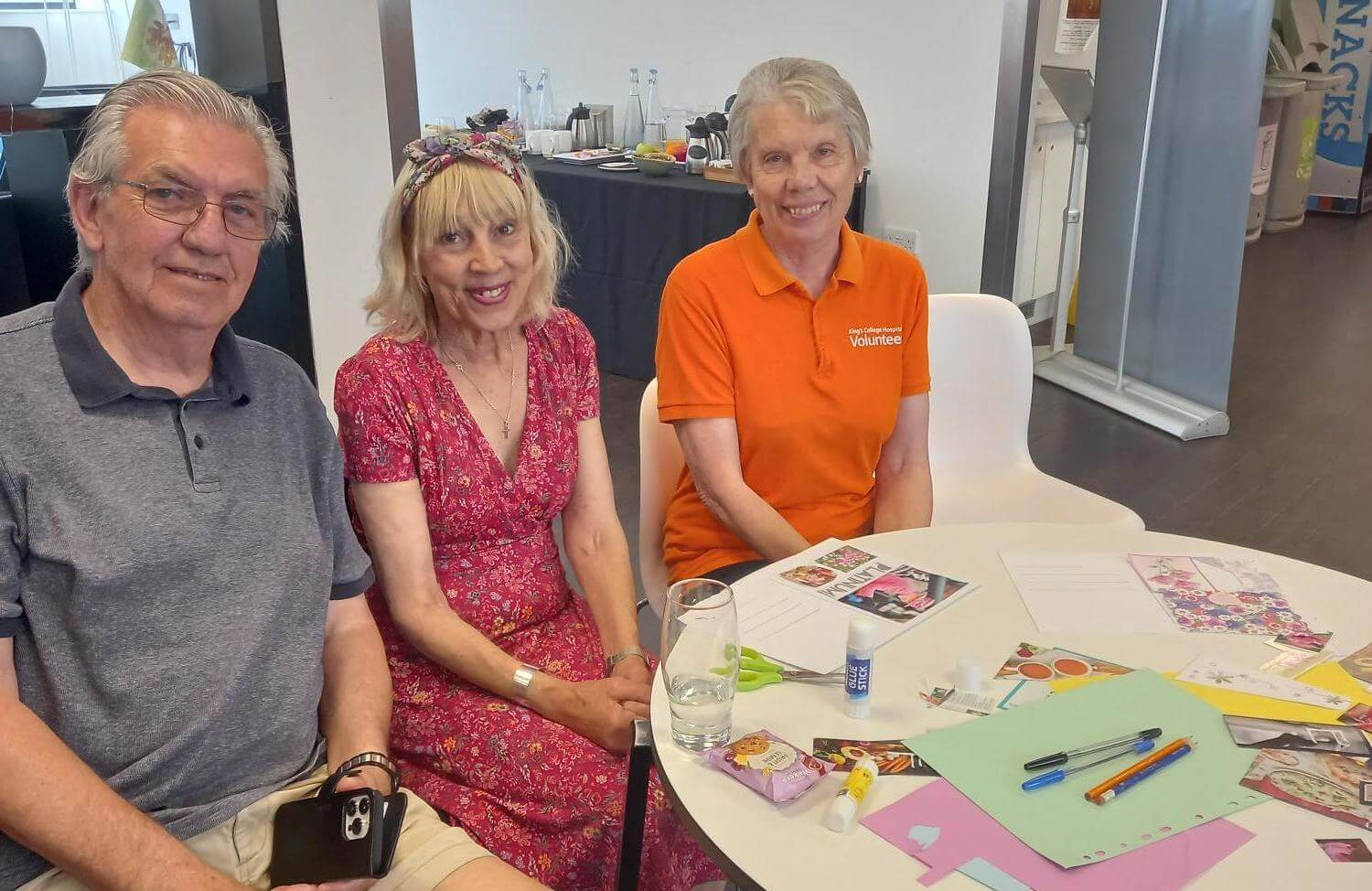 Three seniors seated at a table with craft supplies, smiling at the camera. One of them is wearing an orange volunteer shirt. - Home Instead