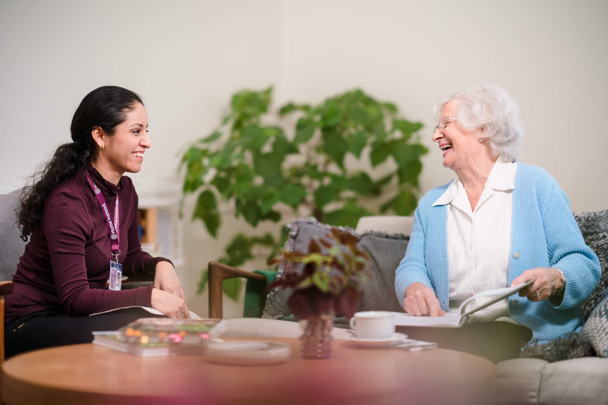 A young woman and an elderly woman smile at each other while sitting on a couch in a cozy living room. - Home Instead