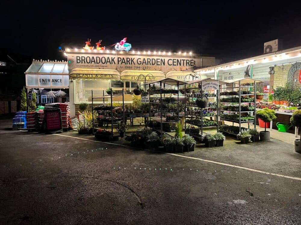 Illuminated garden center at night with potted plants on display and a sign reading "Broadoak Park Garden Centre". - Home Instead