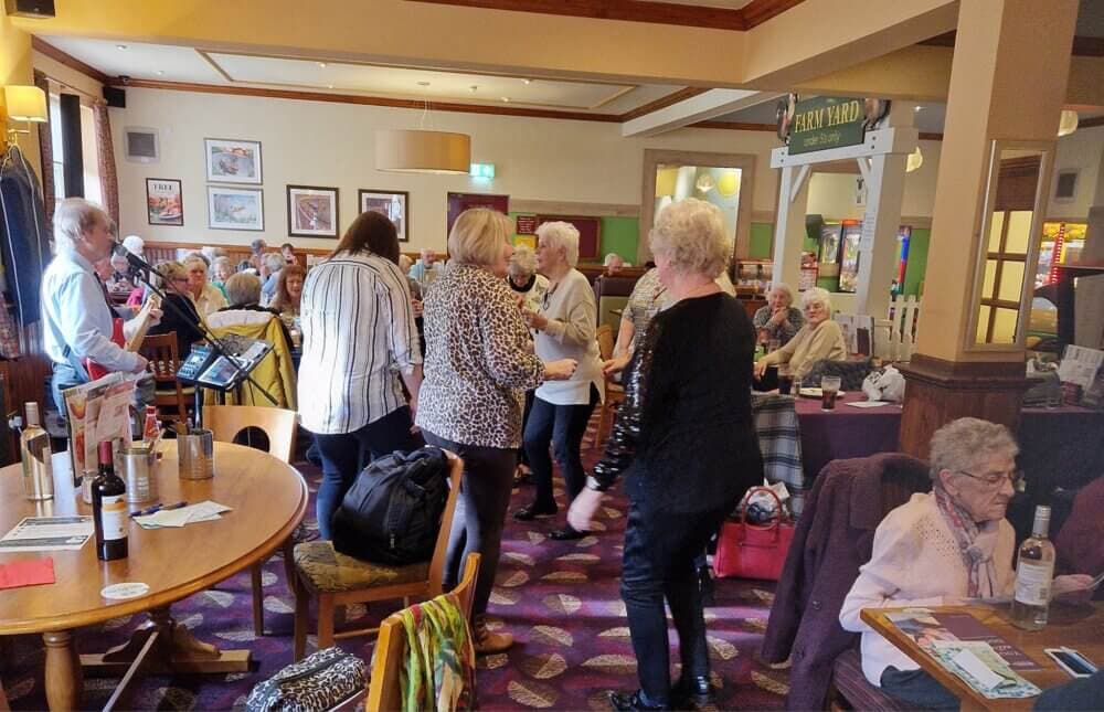A group of elderly people socializing and dancing in a cozy, well-lit room with tables, chairs, and decorative posters. - Home Instead