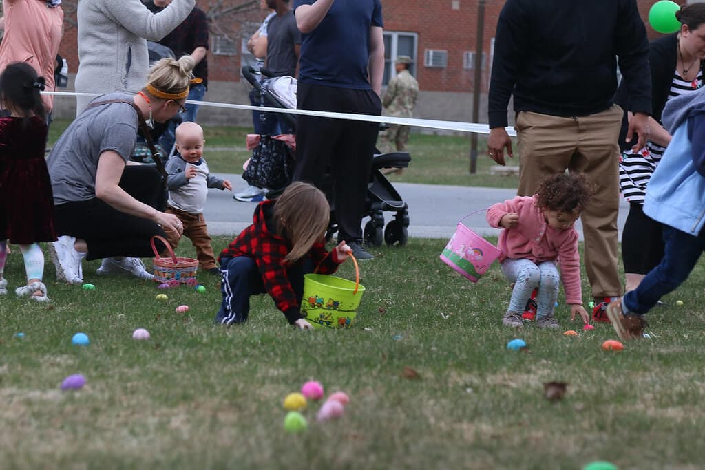 Children and adults participate in an egg hunt, with kids collecting colorful eggs in baskets on a grassy field. - Home Instead