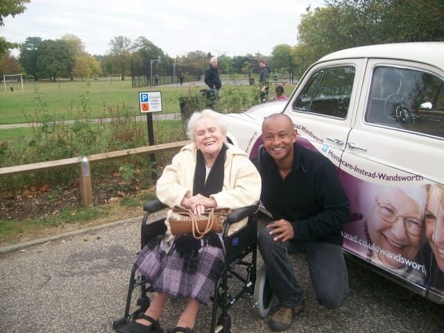 An elderly woman in a wheelchair and a smiling man pose next to a classic white car in a park. - Home Instead