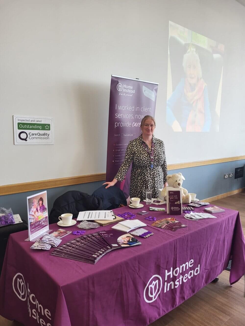 A person stands behind a table with brochures and promotional items for Home Instead, under a banner at a presentation. - Home Instead