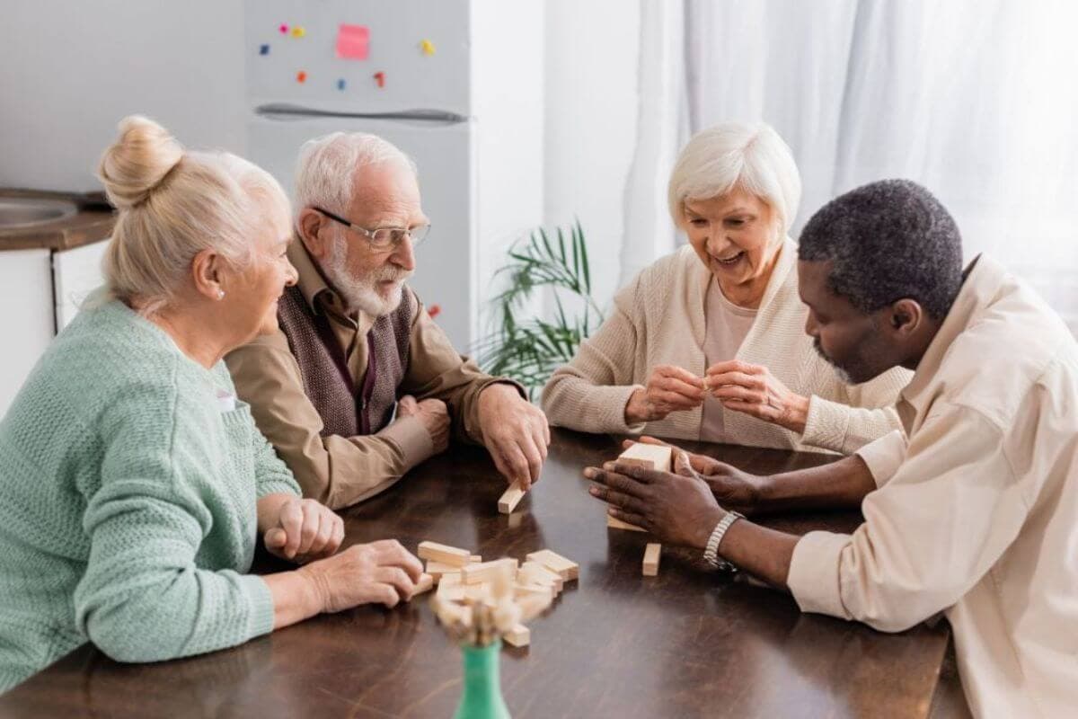 Four elderly people happily playing with wooden blocks at a table in a bright room, sharing laughter and conversation. - Home Instead