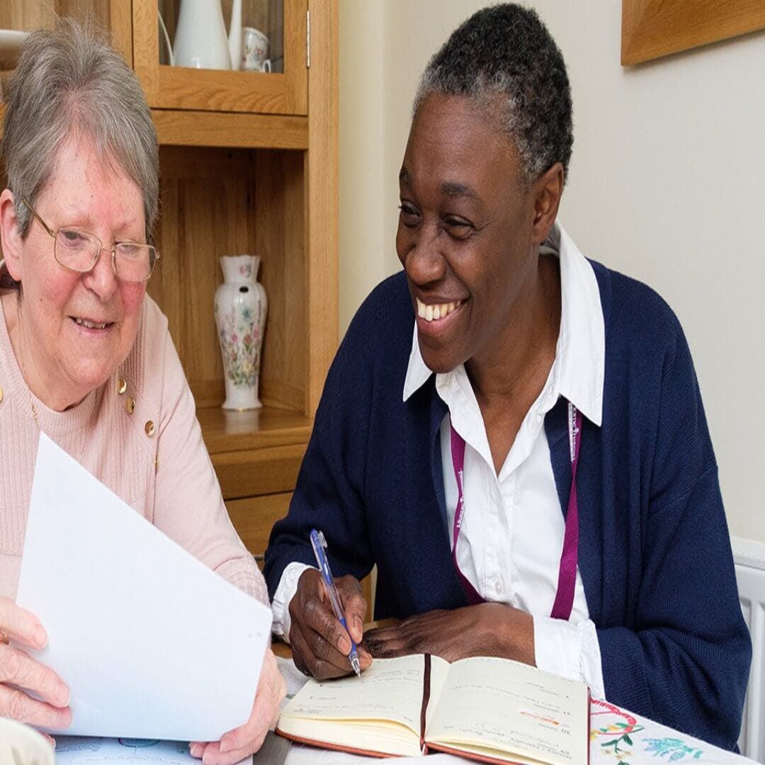 Two women sitting at a table, looking at papers and notebooks, smiling and engaging in a discussion. - Home Instead