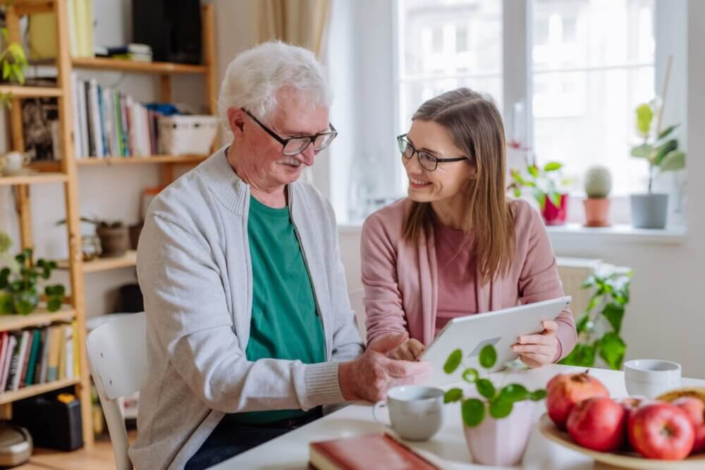 An elderly man and a young woman smile while looking at a tablet in a cosy, well-lit room with plants and bookshelves. - Home Instead Southampton