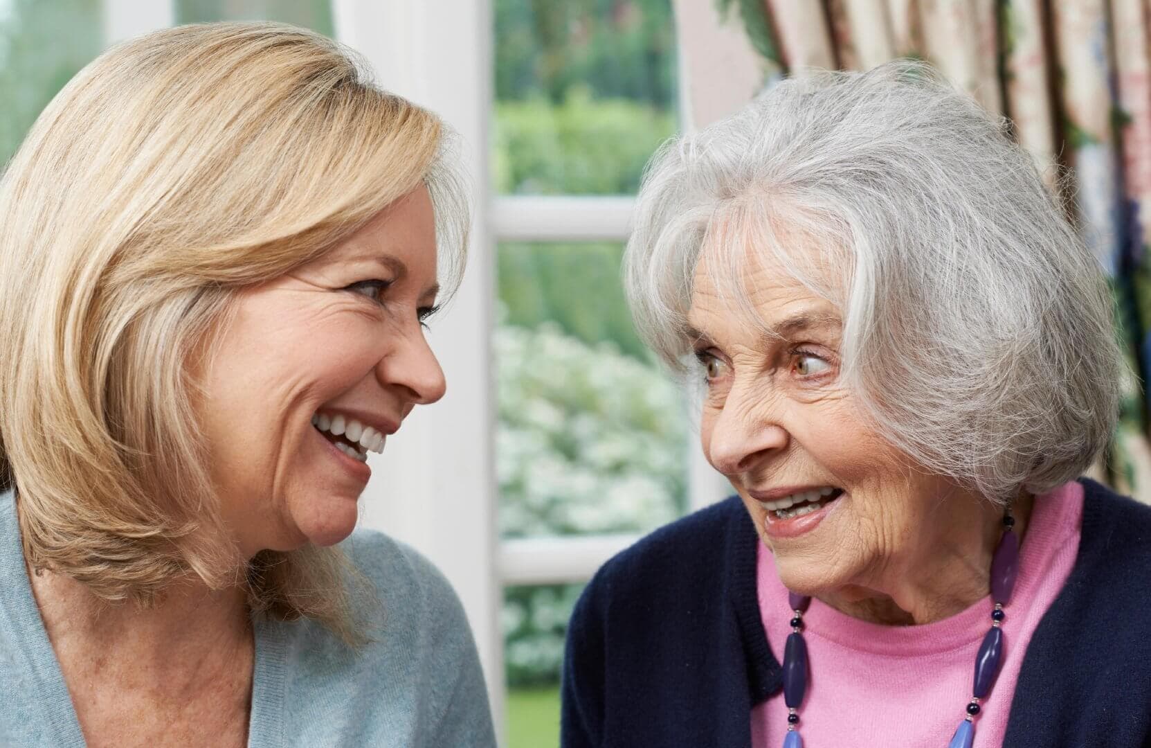 Two women, one younger and one older, smiling at each other in a brightly lit room. - Home Instead