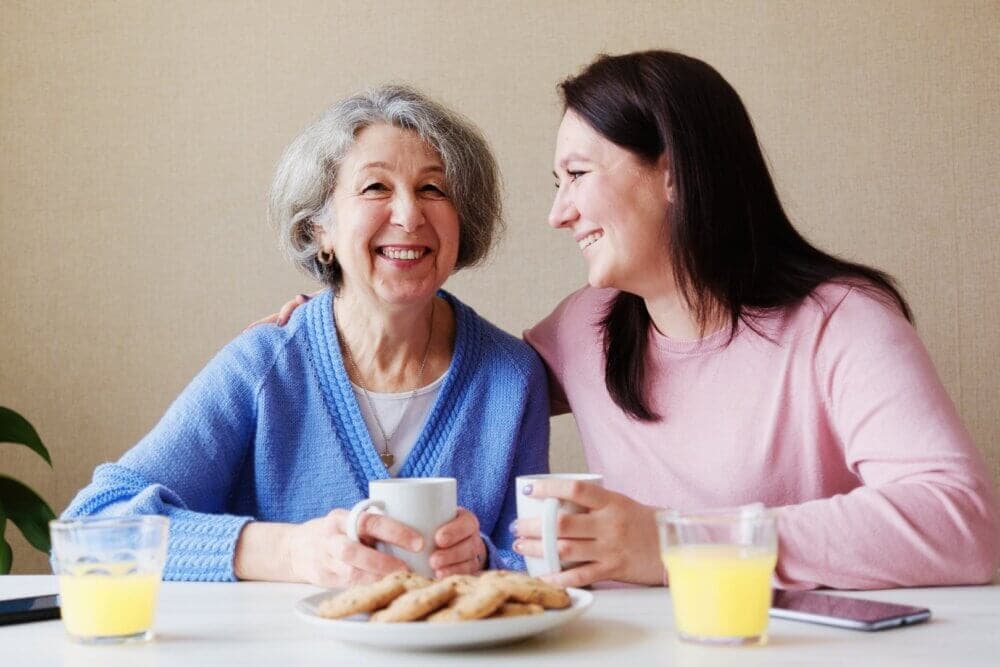 Two women seated at a table with coffee, cookies, and orange juice, smiling and sharing a moment together. - Home Instead Bournemouth & Christchurch