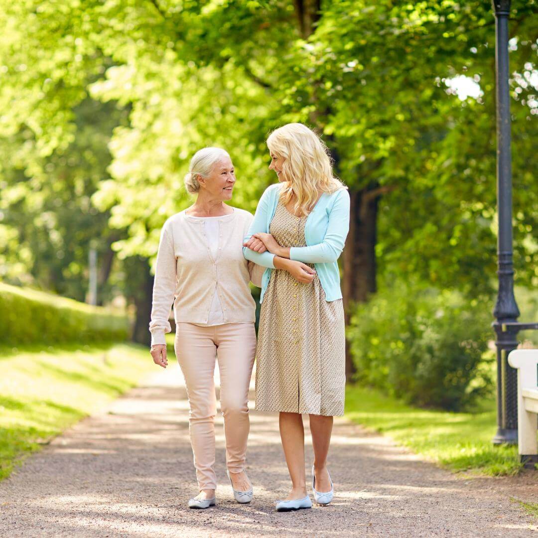 Two women smiling and walking together on a tree-lined path on a sunny day. - Home Instead