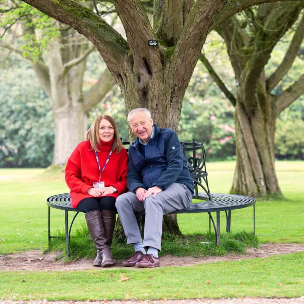 Two people sitting on a metal bench around a large tree trunk in a park, smiling and enjoying nature. - Home Instead