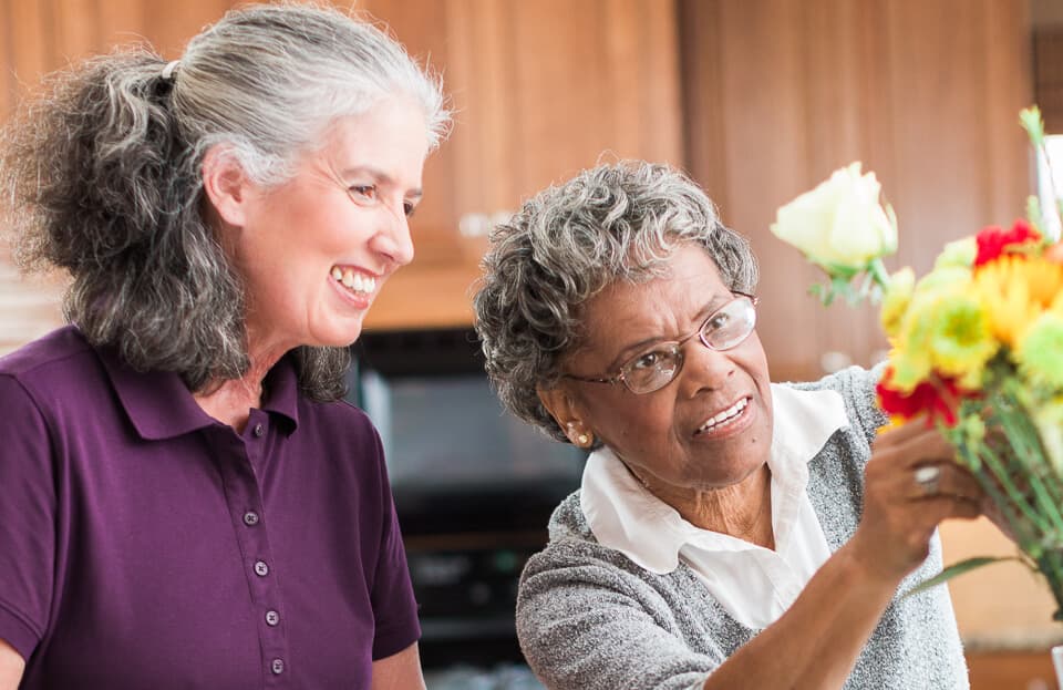 Two women, one older and one younger, arranging flowers and smiling in a warmly lit room. - Home Instead