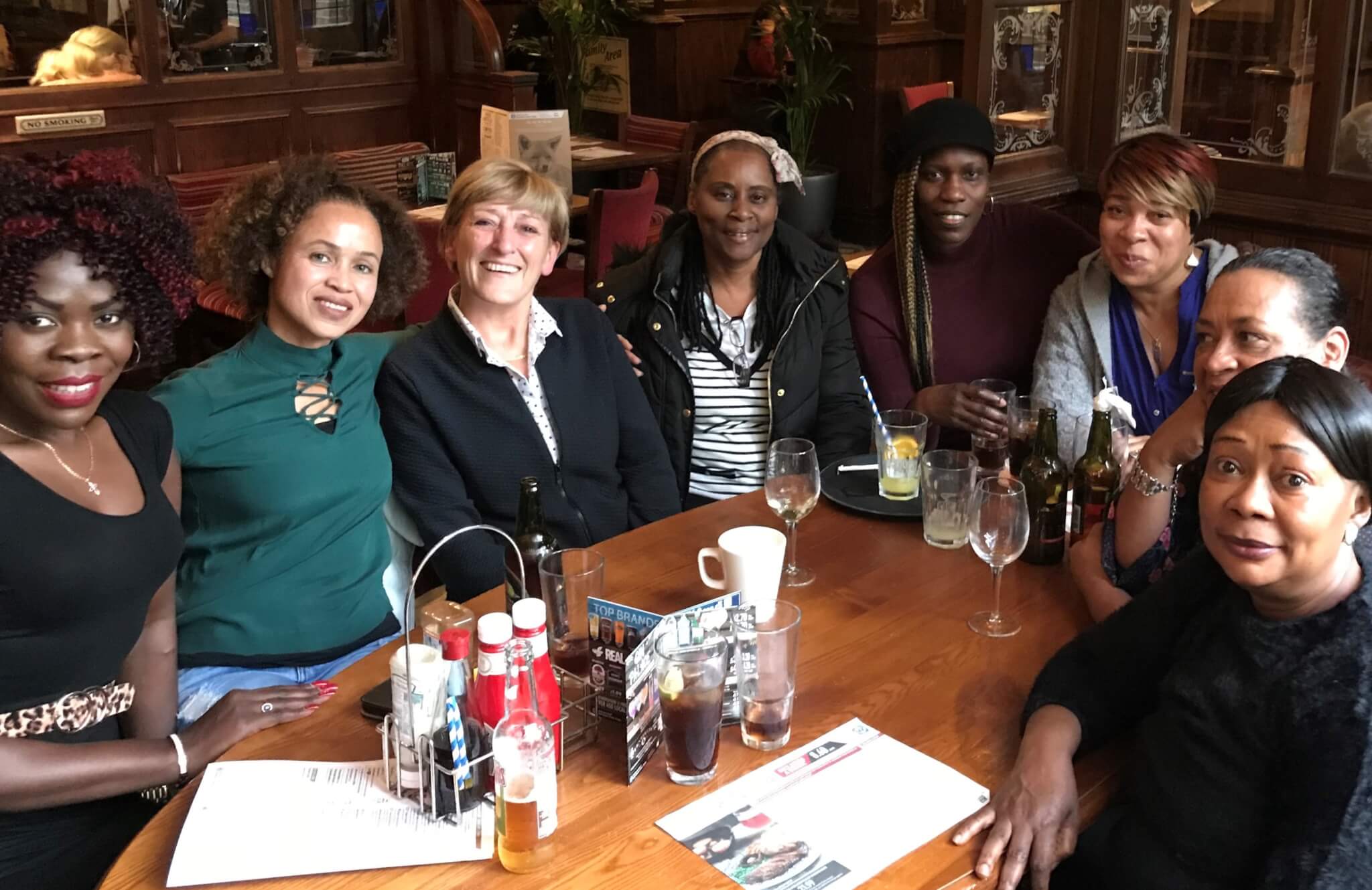 A group of eight women sitting around a wooden table in a restaurant, smiling for the camera. - Home Instead