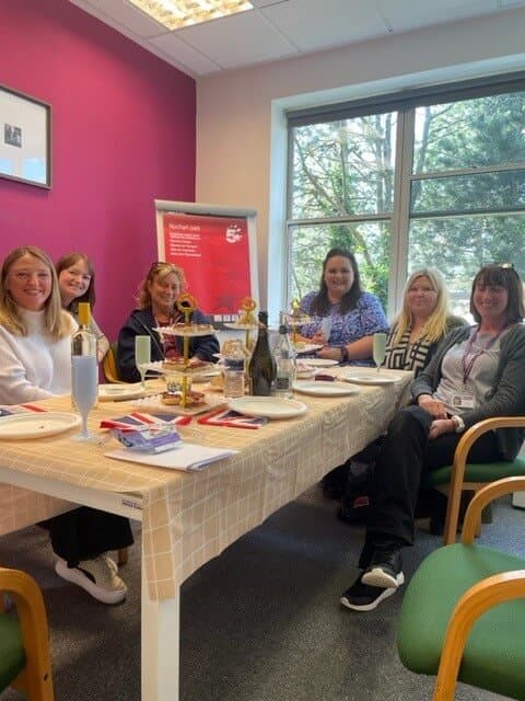 Six people smiling and sitting around a table with food and drinks in a room with a pink wall and large window. - Home Instead