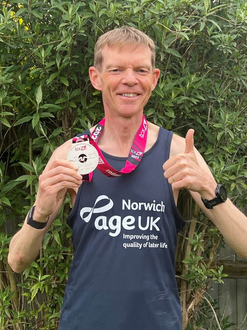 A smiling man holding a medal and giving a thumbs up, wearing a Navy Norwich Age UK shirt, standing outdoors. - Home Instead