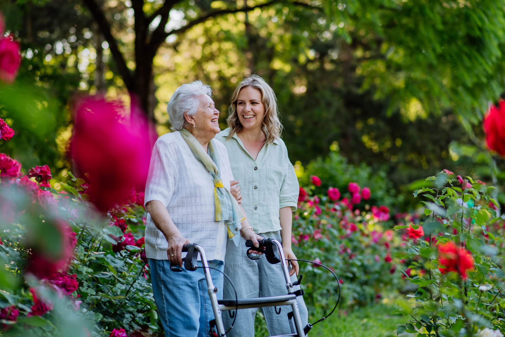 Elderly woman using a walker and young woman smiling together in a garden with blooming flowers. - Home Instead
